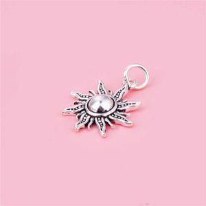 Puede incluir: Un amuleto de plata en forma de sol con una superficie texturizada y un centro redondo. El amuleto tiene un bucle en la parte superior para sujetarlo a una pulsera o collar.