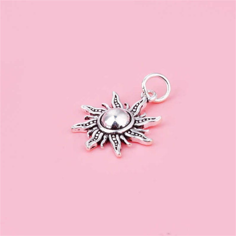 Puede incluir: Un amuleto de plata en forma de sol con una superficie texturizada y un centro redondo. El amuleto tiene un bucle en la parte superior para sujetarlo a una pulsera o collar.