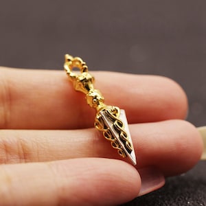 May include: A gold and silver pendant in the shape of a spearhead with a decorative top. The pendant is held in a hand.