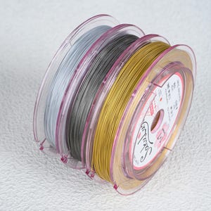 May include: Four spools of beading wire in silver, gray, gold, and light gold. The wire is tightly wound on clear plastic spools with pink accents. The spools are stacked together, with the gold spool in the foreground.