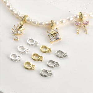 May include: A collection of jewelry findings and charms. The charms include a butterfly, a starburst, and a bow, all with sparkling accents. Several silver and gold-toned lobster clasps are also displayed, along with a pearl necklace.