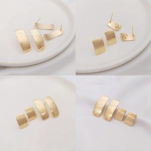 Puede incluir: Pendientes dorados en una variedad de estilos. Los pendientes incluyen tachuelas rectangulares y diseños de barra curvada. El metal tiene una textura cepillada. Los pendientes se muestran sobre una superficie blanca.