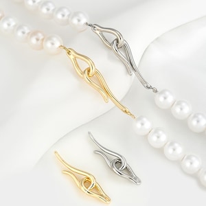 May include: Two pearl necklaces with different clasps. One clasp is gold and the other is silver. Both clasps are in the shape of a knot with a loop at the end.