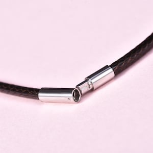 Könnte beinhalten: Nahaufnahme einer dunkelbraunen geflochtenen Lederkette mit einem silberfarbenen Verschluss. Die Kette ist geöffnet und zeigt die beiden getrennten Enden. Der Hintergrund ist ein zartes Rosa.