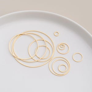 Könnte beinhalten: Eine Sammlung goldfarbener Metall-Creolen in verschiedenen Größen, angeordnet auf einer weißen Oberfläche. Die Reifen reichen von klein bis groß, wobei einige ineinander verschachtelt sind. Das Bild ist gut beleuchtet und hebt den metallischen Glanz des Schmucks hervor.