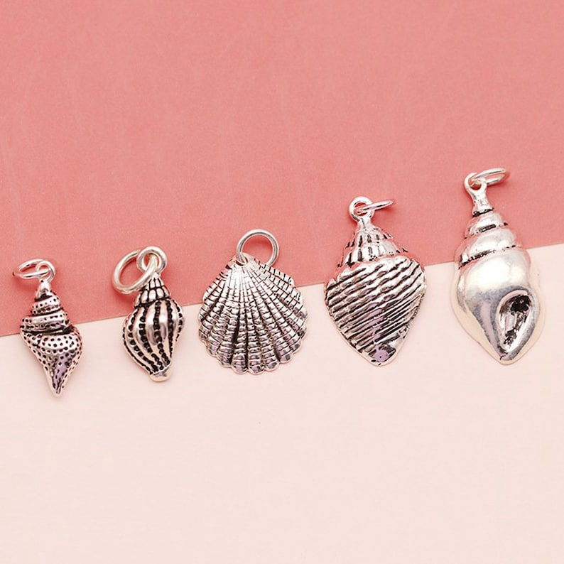 K&ouml;nnte beinhalten: F&uuml;nf silberne Muschel-Anh&auml;nger, jeder mit einem anderen Design. Die Anh&auml;nger sind alle etwa 2,5 cm gro&szlig; und haben eine &Ouml;se oben zum Anbringen an eine Halskette oder ein Armband.