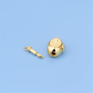 May include: Two gold-colored metal charms on a light blue background. One charm is a rounded shape with two holes, and the other is a cylindrical shape with a pointed end. Both charms have a shiny, reflective surface.