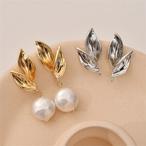 Puede incluir: Un par de pendientes con acabado dorado y plateado. Cada pendiente presenta un delicado diseño de hoja con una perla colgante.