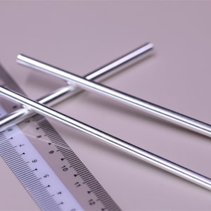 May include: Three silver metal rods are positioned across a ruler. The rods are cylindrical and have a shiny, reflective surface. The ruler is white with black markings, showing measurements in centimetres. The background is a soft, neutral colour.