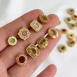 May include: An assortment of gold-coloured jewellery components with diverse gemstone accents. The components showcase intricate designs, including floral and geometric shapes, with gemstones in shades of pink, green, and red. Some pieces are adorned with small, clear crystals.