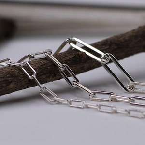 Può includere: Due catene d'argento con maglie rettangolari. Le catene sono appoggiate su un pezzo di legno.