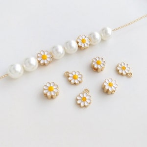 May include: A set of gold-toned daisy charms with white petals and yellow centers. The charms are scattered on a white surface, with a pearl necklace featuring daisy charms in the background.