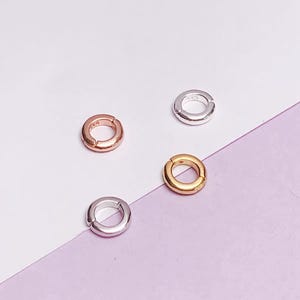 May include: Four small, round jewelry clasps in gold, rose gold, and silver tones are scattered on a white and lavender background. The clasps are open, revealing a small gap. These clasps are used for jewelry making and repairs.