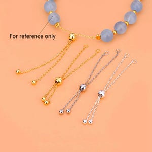 Pode incluir: Uma pulseira com contas esféricas azul-claras separadas por pequenos espaçadores dourados. Inclui correntes de extensão deslizantes ajustáveis em tons de ouro e prata, terminando em pequenas esferas metálicas. Texto: For reference only.