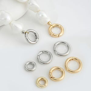 May include: Assortment of gold and silver metal jump rings. The jump rings are different sizes and are arranged on a white surface.