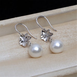 Puede incluir: Un par de pendientes de plata con una perla blanca y un pequeño diseño floral.