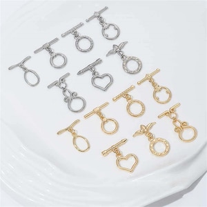 Może przedstawiać: Asortyment srebrnych i złotych zapięć typu toggle w różnych wzorach, w tym okręgi, serca i kształty dekoracyjne. Zapięcia są ułożone na białej, lekko teksturowanej powierzchni.