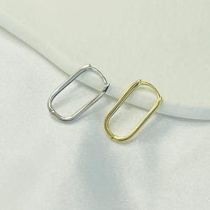 Könnte beinhalten: Zwei ovale Creolen, eine silberne und eine goldene, werden auf einer weißen Oberfläche gezeigt. Die Ohrringe haben ein schlichtes, minimalistisches Design mit glatter, polierter Oberfläche. Der goldene Ohrring hat einen warmen, hellen Ton.