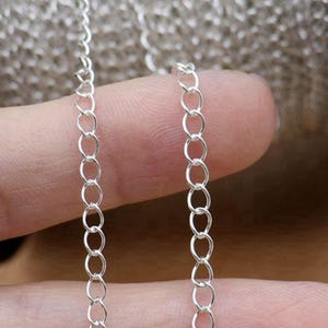 May include: Close-up of a delicate silver chain. The chain is made up of small, evenly spaced oval links. The links are bright and reflective, suggesting a polished finish. The chain is held against a blurred background, highlighting its intricate design.