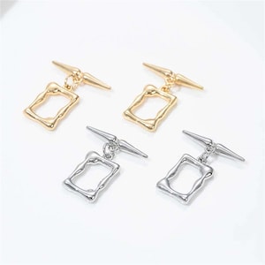 Op de afbeelding: Vier paar oorbellen, twee goudkleurig en twee zilverkleurig. Elke oorbel heeft een rechthoekige, open-frame hanger die aan een T-vormige pin hangt. De hangers hebben een onregelmatig, getextureerd oppervlak.