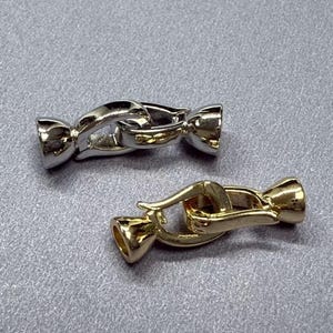 May include: Two metal jewelry clasps. One is silver-colored, the other is gold-colored. Both clasps have a curved design with a hook and eye closure. The clasps are designed to connect two pieces of jewelry.