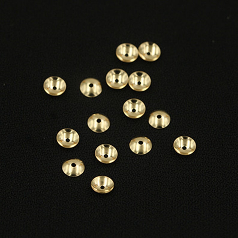 May include: A group of 15 gold-colored metal bead caps scattered on a black surface. The caps are round with a flat top and a small hole in the center.