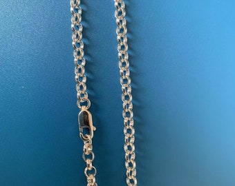 May include: A silver-colored chain necklace with a lobster clasp, displayed against a solid blue background. The chain features a series of interconnected, rounded links. The clasp is visible at the top left, and the chain hangs vertically.