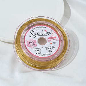 May include: A spool of gold-colored nylon-coated wire, labeled "SakuLux," with details including 0.40 mm coated out diameter and 100m length. The label also indicates "Stainless Steel" and "Made in Japan."