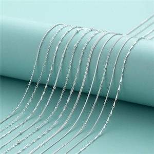Può includere: Una collezione di collane d'argento con vari stili di catena, tra cui catene a perline, intrecciate e a scatola. Le collane sono esposte su uno sfondo verde acqua chiaro, mostrando i loro diversi design e texture. Le catene sono delicate e lucenti.