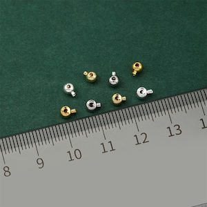 Puede incluir: Un conjunto de pequeñas cuentas metálicas redondas en tonos dorados y plateados. Las cuentas tienen un pequeño lazo. Una regla con marcas de centímetros está presente, indicando el tamaño de las cuentas.