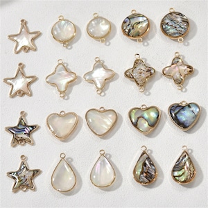 May include: Assortment of 20 gold-toned metal charms with various shapes and designs. The charms are made of white and iridescent mother-of-pearl. The shapes include stars, circles, hearts, teardrops, and crosses.