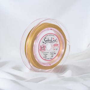 May include: A spool of gold-colored nylon-coated wire, labeled "SakuLux", is displayed. The wire is wound around a clear plastic spool. The label indicates the wire is 0.60 mm in diameter and 100 m in length. The text "Stainless Steel" is also visible.