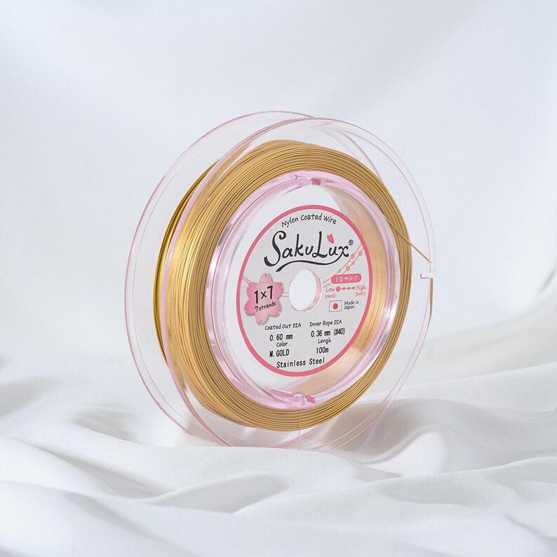 May include: A spool of gold-colored nylon-coated wire, labeled "SakuLux", is displayed. The wire is wound around a clear plastic spool. The label indicates the wire is 0.60 mm in diameter and 100 m in length. The text "Stainless Steel" is also visible.