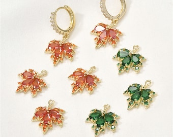 May include: A set of six gold-tone earrings featuring a maple leaf design with red and green crystal accents. The earrings are available as individual charms or as hoop earrings.