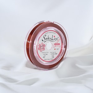 May include: A spool of red nylon-coated wire, labeled "SakuLux," is displayed against a white background. The label indicates the wire is 0.40 mm in diameter and 100 m in length. The spool is made of stainless steel.