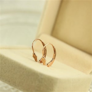Pode incluir: Um par de brincos de ouro rosa com fecho de alavanca. Os brincos são exibidos dentro de uma caixa de joias bege. Os brincos têm um design simples e elegante, com um aro curvo e um fecho de alavanca.