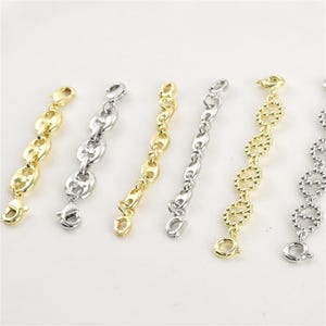 May include: Close-up of several gold and silver-colored chain extenders with lobster claw clasps. The extenders feature various link designs, including oval and bean-shaped links. The jewelry findings are displayed against a white background.