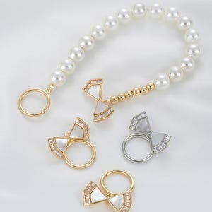 May include: An assortment of jewelry pieces, including rings and a bracelet, displayed on a white surface. The rings feature a bow-tie design with a mother-of-pearl inlay and sparkling accents. The bracelet is made of white pearls and gold-toned metal.