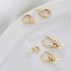 Puede incluir: Pendientes dorados con una variedad de diseños. Algunos presentan un lazo simple, mientras que otros tienen una perla colgante. Los pendientes se exhiben sobre una superficie blanca, mostrando su acabado pulido y estilo elegante.