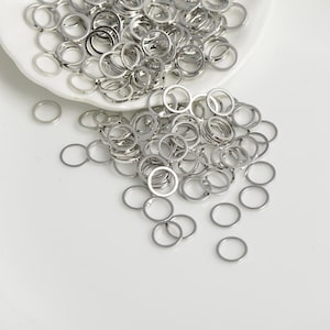 May include: A collection of small, silver-coloured metal rings scattered on a white surface and in a white dish. The rings are uniform in size and shape, with a circular design. The image is well-lit, highlighting the metallic sheen of the rings.