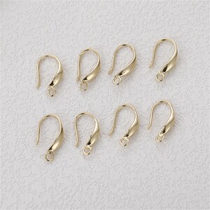 May include: Eight gold-colored earring hooks with a curved design. Each hook has a small loop at the bottom for attaching charms or beads. The hooks are displayed on a light-colored textured surface.