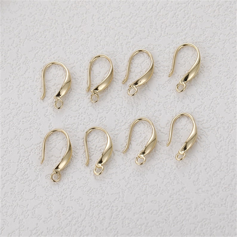 May include: Eight gold-colored earring hooks with a curved design. Each hook has a small loop at the bottom for attaching charms or beads. The hooks are displayed on a light-colored textured surface.