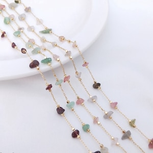 May include: Five gold chains with small, colourful gemstone beads. The chains are layered and arranged on a white surface.
