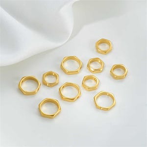 May include: A collection of ten gold-toned metal hexagonal jump rings. The rings are of varying sizes and are arranged on a white surface.