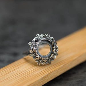 Puede incluir: Un anillo de plata con un diseño floral. El anillo presenta un engaste circular rodeado de pequeñas flores detalladas. El anillo se muestra sobre una superficie de madera, con un fondo oscuro.