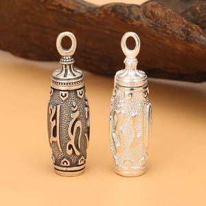 Puede incluir: Dos colgantes de plata con diseños intrincados. Los colgantes son cilíndricos y presentan letras y patrones en relieve. Los colgantes miden aproximadamente 5 cm de largo.