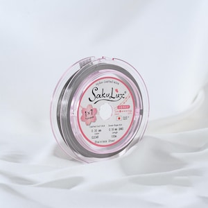 May include: A clear, nylon-coated wire spool with the brand name "SakuLux" printed on the label. The label also indicates the wire is 0.30 mm in diameter and 100m in length. The spool is encased in a clear, pink plastic container.