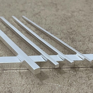 May include: A close-up shot of several silver metal bars. The bars are rectangular and of varying lengths. They appear to be made of a shiny, reflective material. The bars are arranged on a neutral-toned surface, creating a minimalist aesthetic.