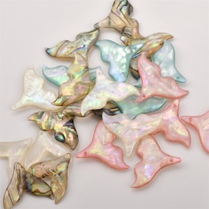 May include: A collection of whale tail-shaped charms crafted from iridescent shell material. The pieces feature a variety of colours including soft pink, pale blue, white, and multi-coloured pearlescent tones with a smooth, polished finish.
