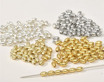 May include: A collection of oval-shaped metal beads in gold and silver colors. The beads are scattered on a white surface, with some arranged in a line on a thin needle.